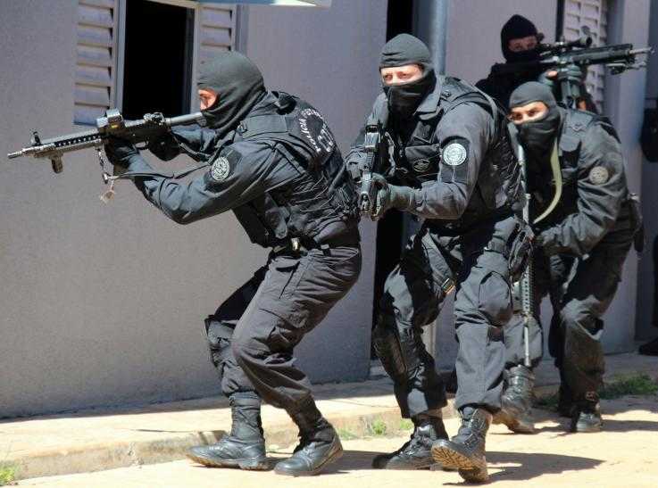 Four police officers wearing balaclavas and pointing in several directions with their machine guns creep round the edge of a building