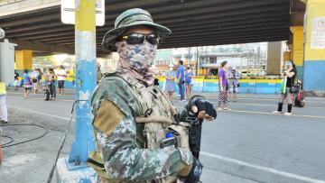 A soldier stands in a street with a gun, wearing a face mask.