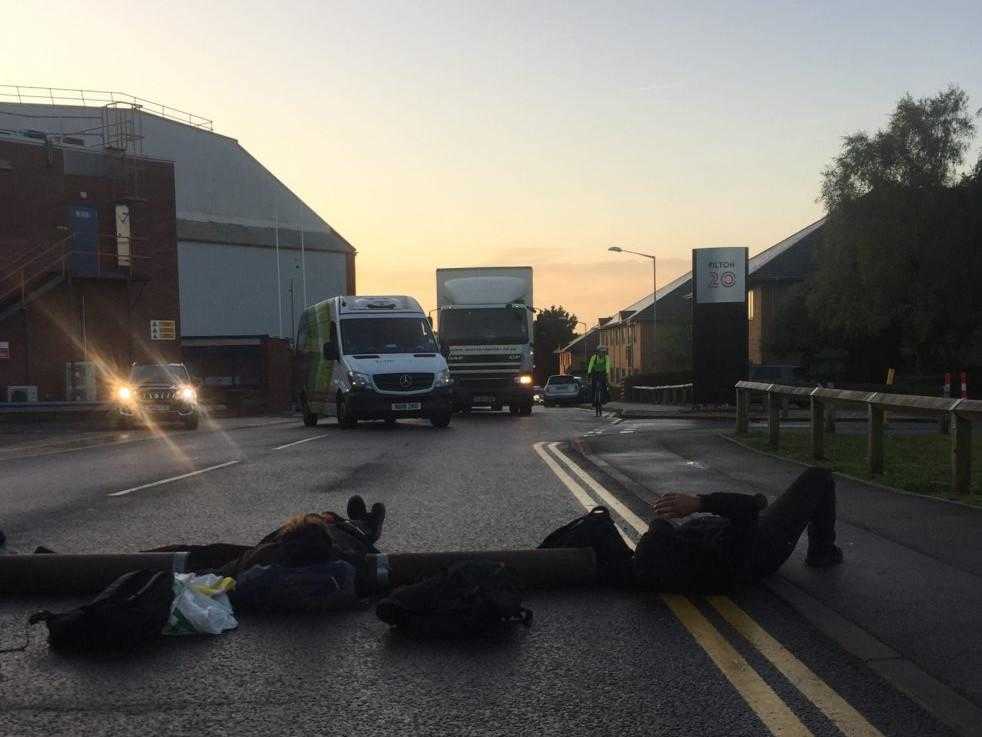 A photo taken at dawn. In the foreground a number of people are lying on the road attached together with metal tubes. In the distance are a number of vehicles on the road.