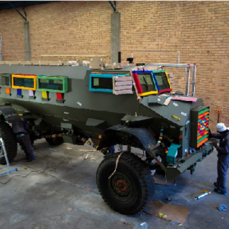A largely undecorated Casspir stands in a warehouse with brick walls and a concrete floor.  The khaki paintwork of the Casspir is interrupted by bright, multicoloured beading on the window frames and grille.  An artist in overalls is working on the beading on the grille and near the back wheel there is a platform ladder.