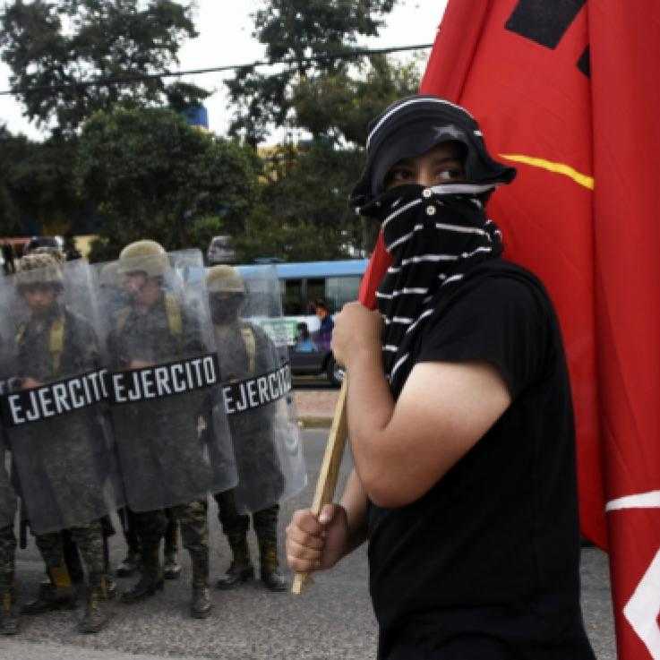 An activist with his face covered holds a red flag. He is stood in front of a line of police officers and military personnel holding shields.