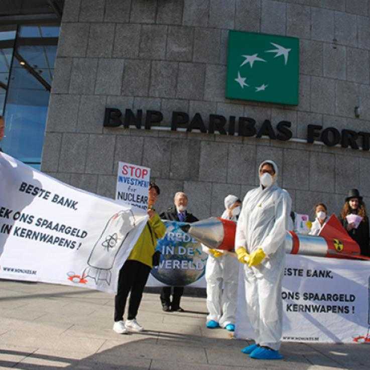 Activists dressed in white "bio hazard suits" and wearing gas masks hold banners reading "don't invest in nuclear weapons" outside a building with the BNP Paribas logo