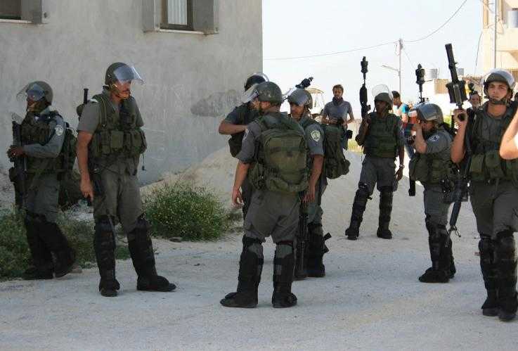 MAGAV border guards dressed in body armour and helmets stand at ease on a street corner.  Some have their guns down by their side and others have them pointing up in the air.