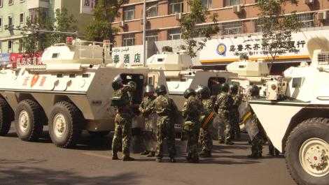 Cuatro vehículos que parecen tanques permanecen inmóviles en una calle. La policía vestida con uniforme de combate con cascos y escudos antidisturbios se yergue casualmente en el camino que los separa.