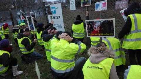 Several dozen protesters wearing yellow vests kneel on the grass outside a fence. On the fence is the sign of the company, Alsetex.  Several protesters are holding signs depicting injuries caused by the companies products
