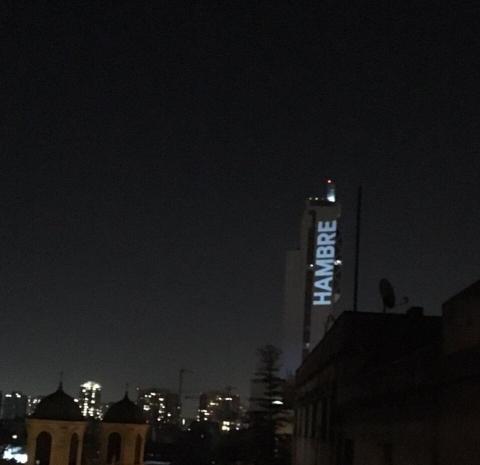 The word "hambre" ("hunger" in Spanish) is projected in huge letters onto a building 