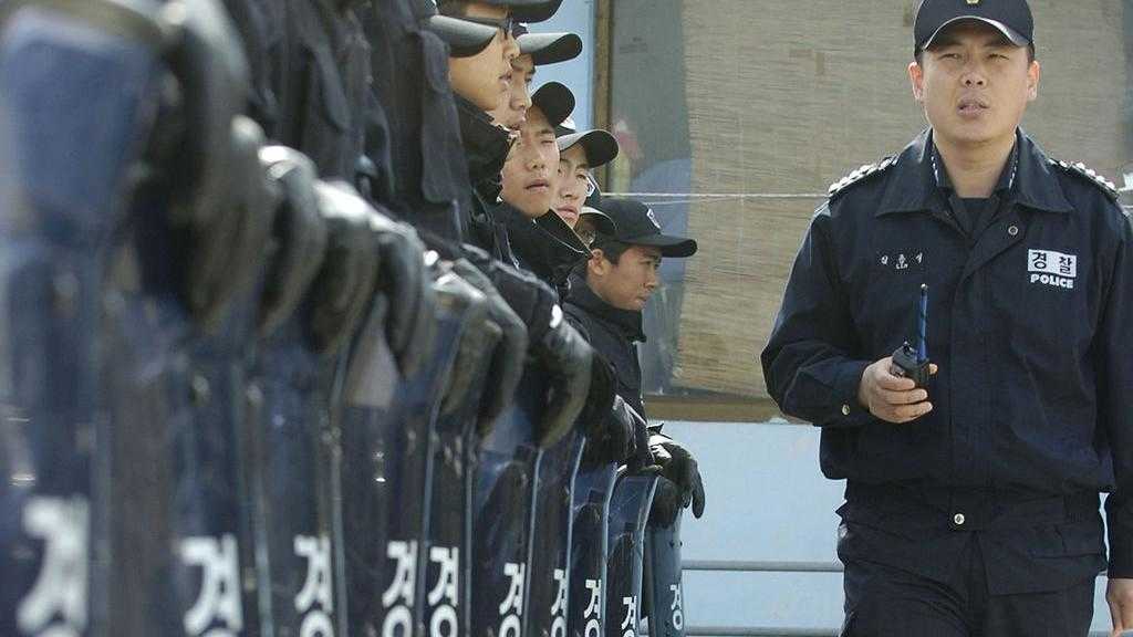 An officer with a walkie-talkie inspects a line of police conscripts wearing blue uniforms and baseball caps and resting their leather-gloved hands on transparent riot shields emblazoned with the word 'police' in Korean script