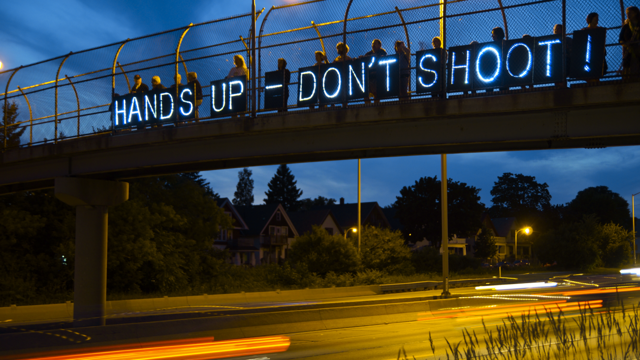 Aktivisten halten Transparente von einer Brücke, die nachts mit LED beleuchtet sind, mit dem Text: “Hände hoch – nicht schießen”. Quelle: flickr