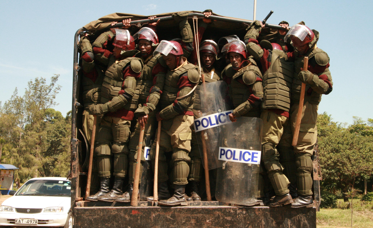 A large number of police armed with clubs wait to jump out of a van in Kenya. The police are wearing a lot of armour, shields, helmets and clubs.
