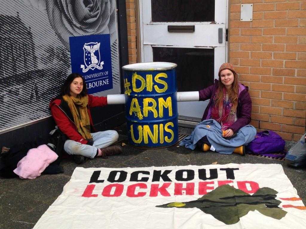 Two student activists lock on to a barrel filled with concrete. The barrel is painted blue and has the words "Disarm Unis" painted on it. In front of the activists there is a banner reading "Lockout Lockheed". Behind them is a sign for the University of Melbourne