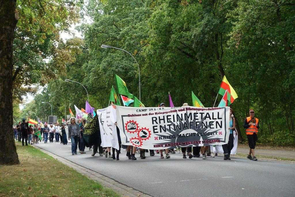 A large group of people march down a street. The front group are holding a banner reading "disarm Rheinmetall" in German