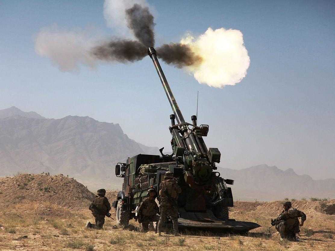 A large cannon is fired in desert terrain. A number of soldiers stand around the side.