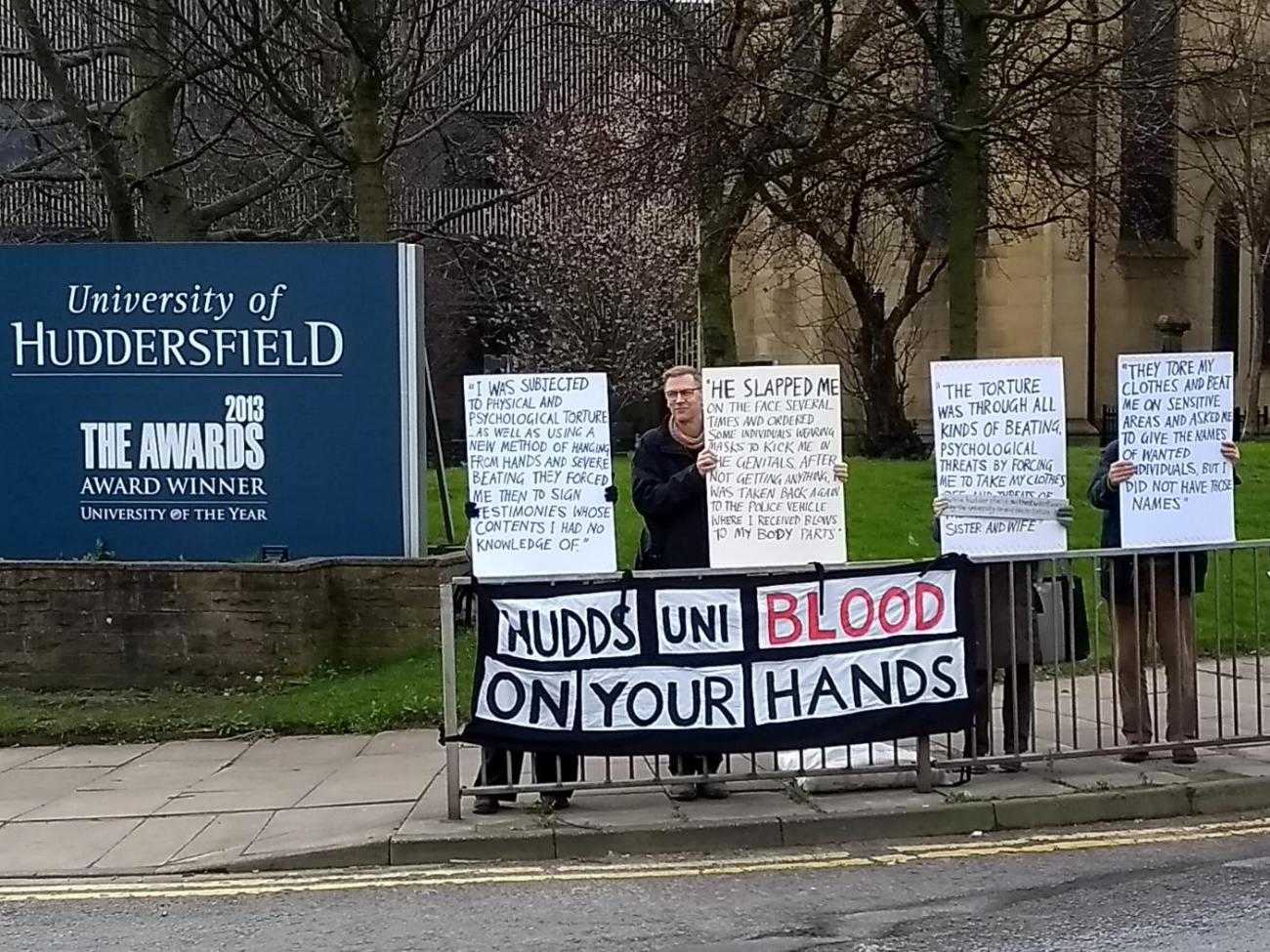 Four protesters hold signs outside the University of Huddersfield. The placards detail human rights abuses at the Bahrain Royal Academy