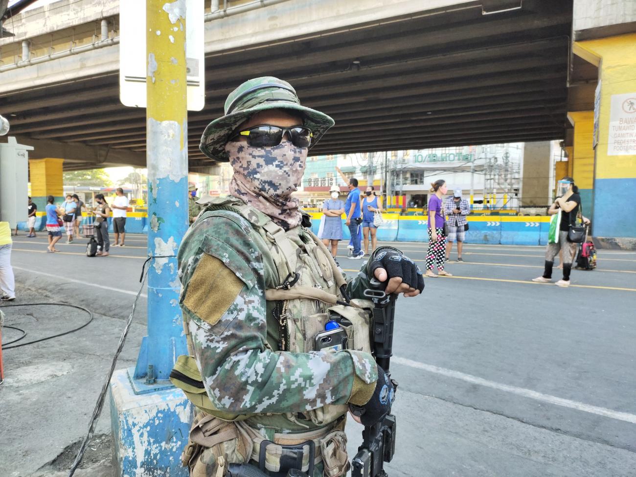 Un soldado está parado en una calle con un arma, usando un tapabocas