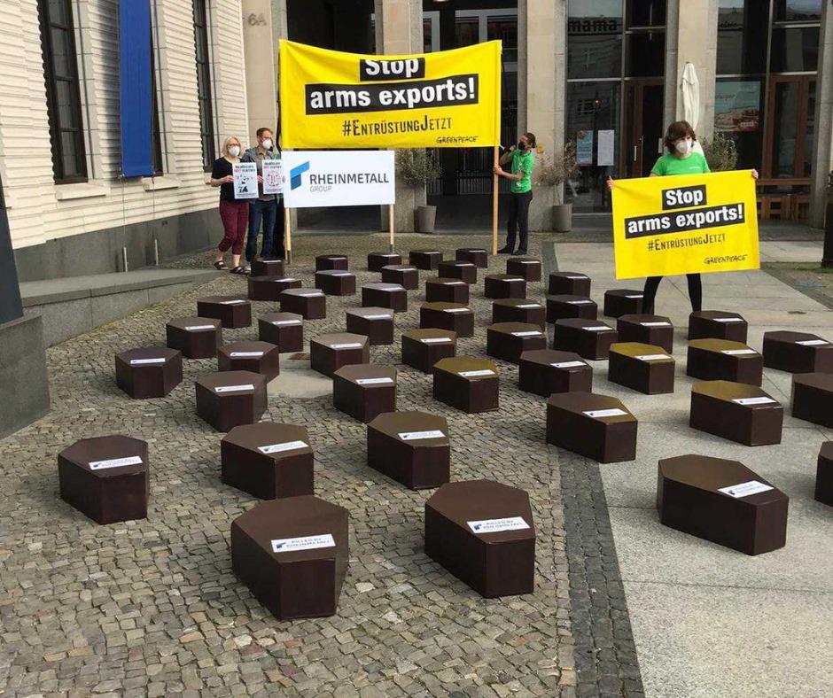 In the foreground are dozens of small coffins. In the background people stand with yellow banners