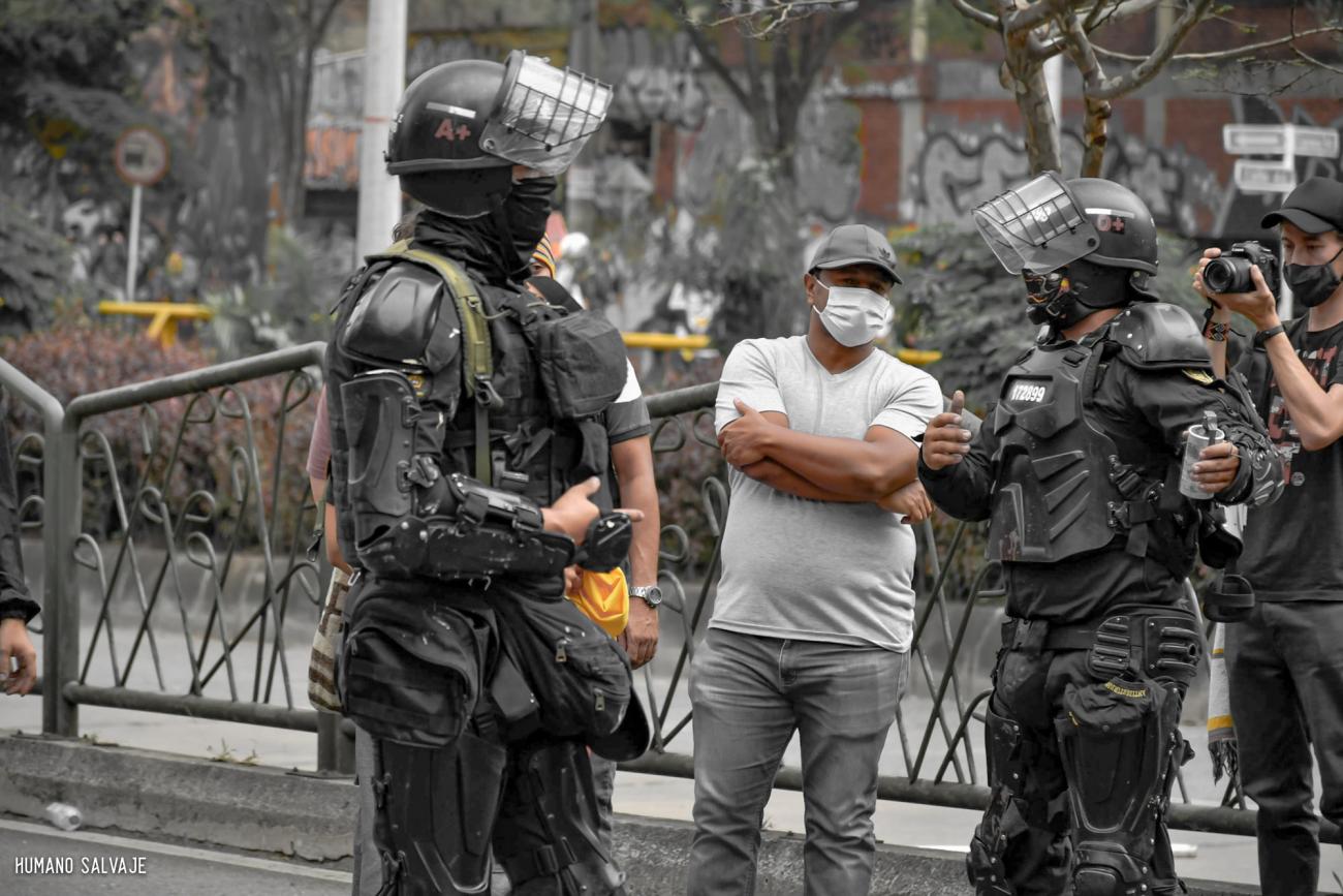 Varios policías fuertemente blindados se sitúan alrededor de un hombre vestido de manera informal. Uno de los policías sostiene una granada