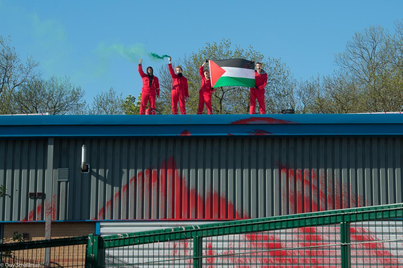 Four people stand on the roof of a metal building behind a fence. They are wearing red and holding a palestine flag. The building has red paint on it.