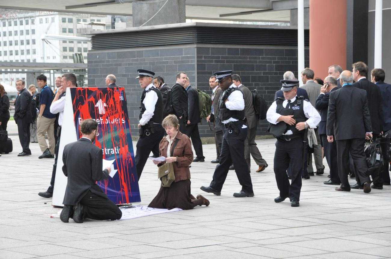 Two activists kneel in front of a sign directing people to DSEI, which has been covered in red paint, to symbolise spilt blood. Behind them a queue of people wait to go into the fair. Three police officers are approaching the activists.