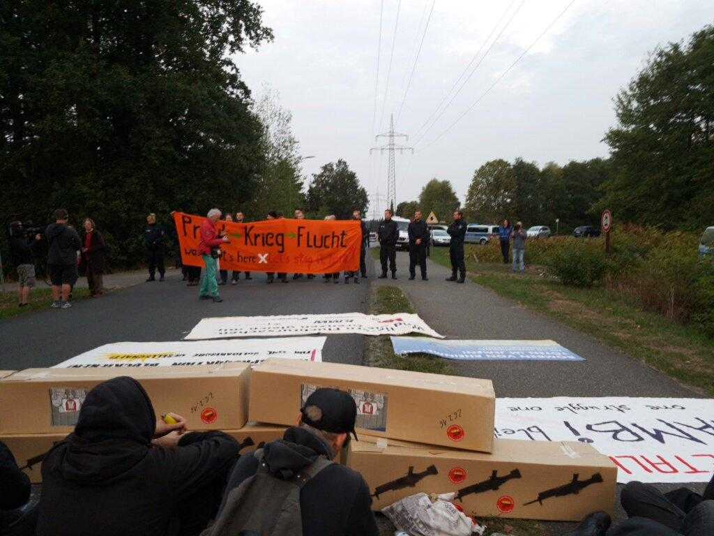 Activists sit behind a blockade of "delivery boxes" across the road. In the distance there is an orange banner reading "war starts here, let's stop it here!"