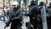 Heavily armed police officers in the UK face protesters. One is carrying a large weapon, another a shield.