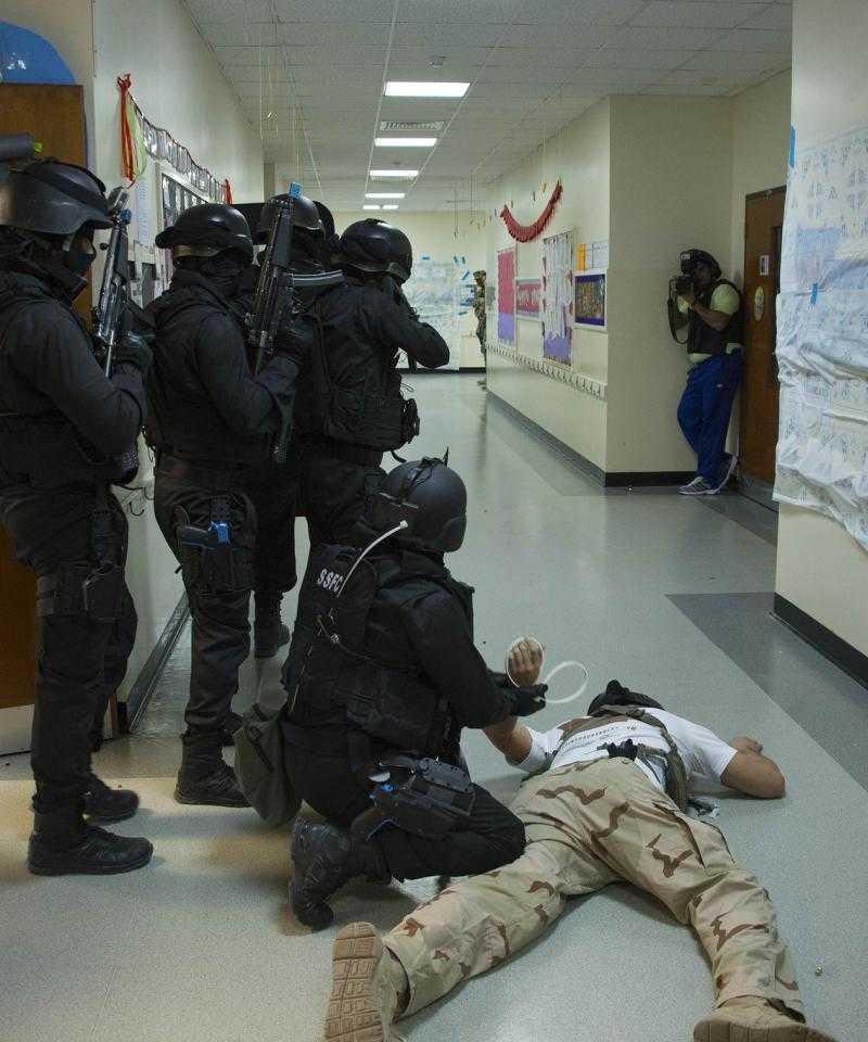 A training exercise involving Bahraini and US police. A man being "arrested" is held to the floor while others point guns down a corridor. There is a photographer in the background.