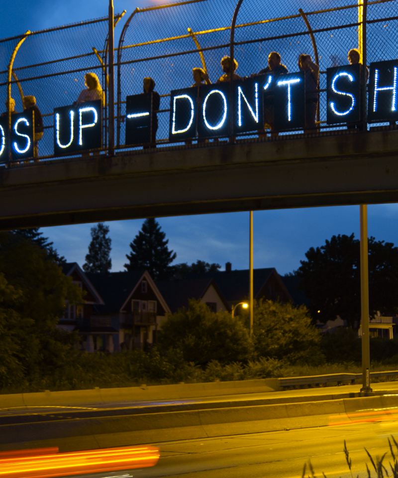 Protesters hold banners with small lights reading "Hands up don't shoot" on a bridge over a road at dusk