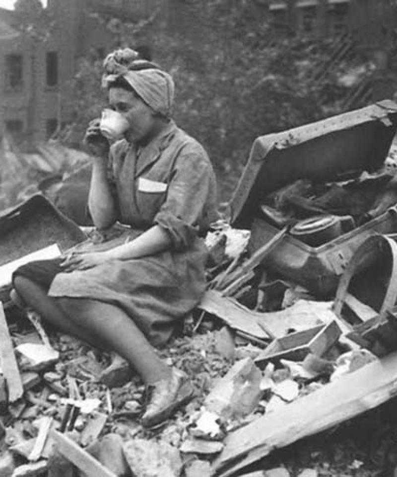 A woman drinks a cup of tea amid a destroyed building. The photo is black and white.