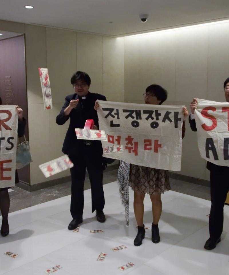Quatre personnes brandissant des pancartes disant «Stop ADEX» en anglais et en coréen. L'une des personnes porte un collier de chien.