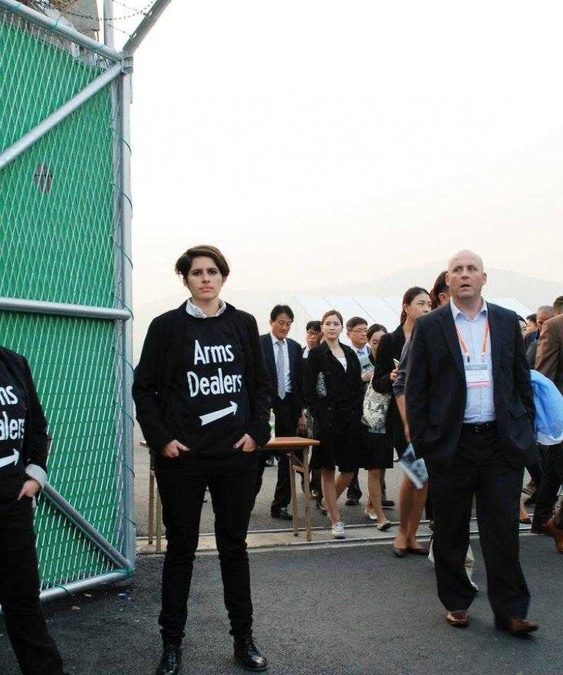 Hannah and another protester wearing t-shirts with the words "arms dealer" with an arrow. The arrow points to a line of people, mainly men in suits, leaving the ADEX arms fair.
