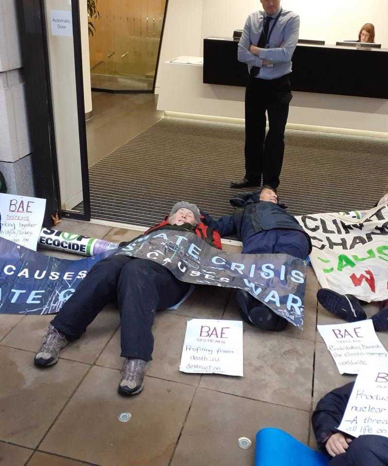 A number of people lie on the ground outside an office door. In the background there is a security guard stood still.