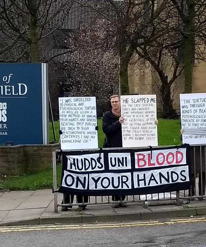 Four protesters hold signs outside the University of Huddersfield. The placards detail human rights abuses at the Bahrain Royal Academy
