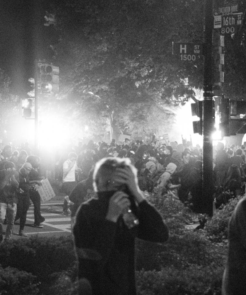 A black and white photo at night, showing large numbers of people walking through smoke with their mouths covered