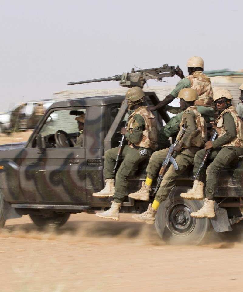 A group of soldiers on a large jeep driving through the desert. The jeep has a large gun on top and the soldiers are armed.