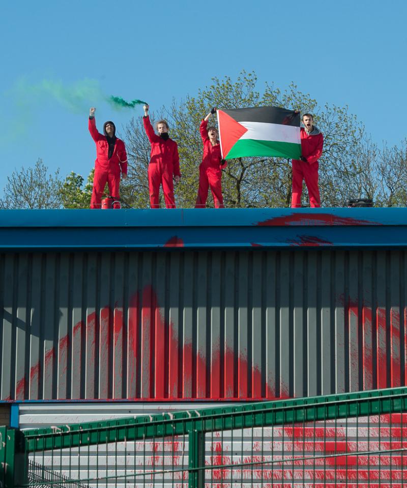 Four people stand on the roof of a metal building behind a fence. They are wearing red and holding a palestine flag. The building has red paint on it.
