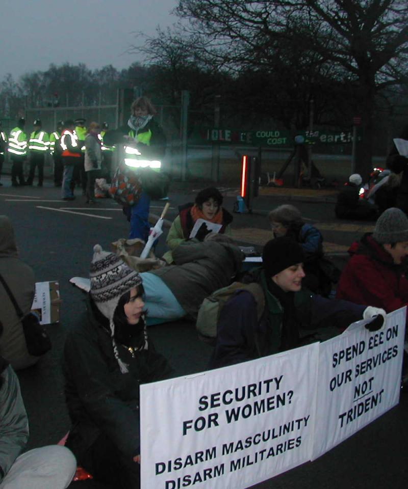 Women sit across a road in a protest against nuclear weapons. They are holding signs