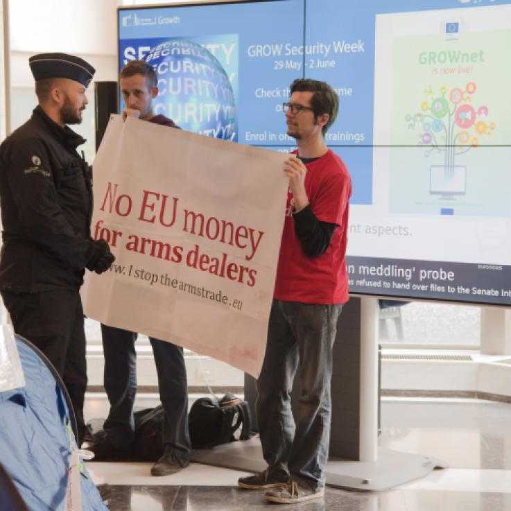 Protesters holding a sign reading 'No EU money for arms dealers' talking to police officers in the foyer of the European Commission