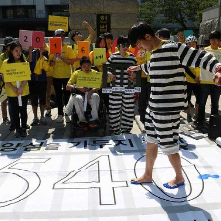 Protests in Seoul against the imprisonment of conscientious objectors; a man dressed as a prisoner walks across across a banner with 540 written on (the number of COs then in jail)