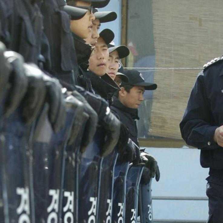 An officer with a walkie-talkie inspects a line of police conscripts wearing blue uniforms and baseball caps and resting their leather-gloved hands on transparent riot shields emblazoned with the word 'police' in Korean script