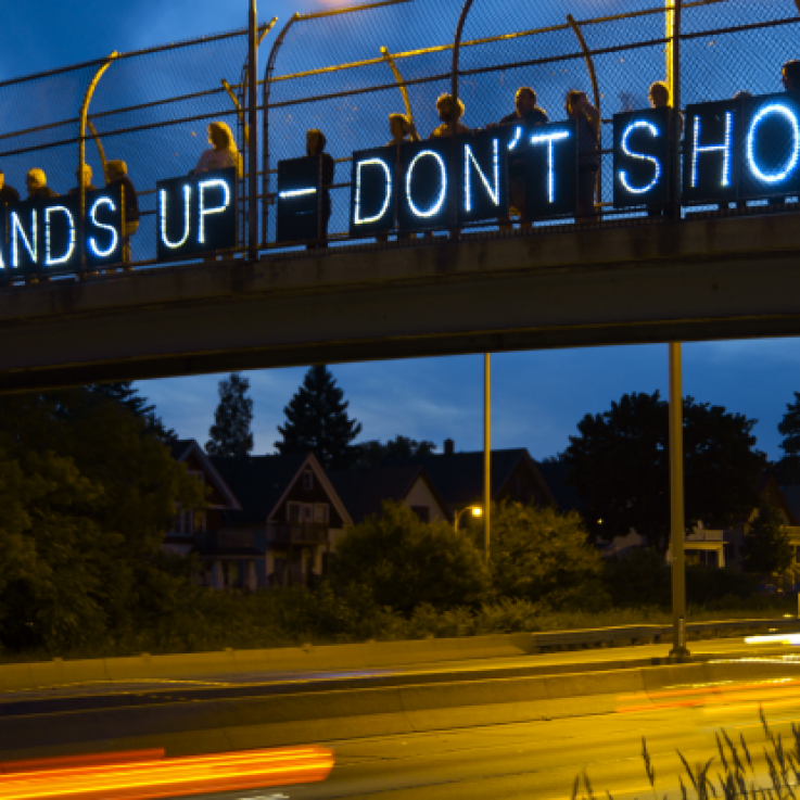 Protesters hold banners with small lights reading "Hands up don't shoot" on a bridge over a road at dusk