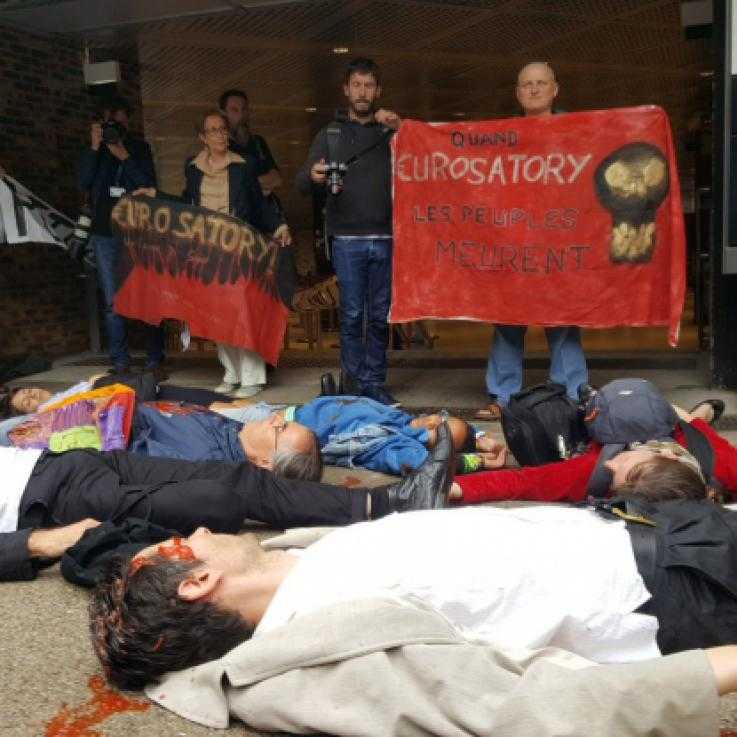 A large group of people lie on the floor as part of a "die-in". In the background other protesters stand with banners.
