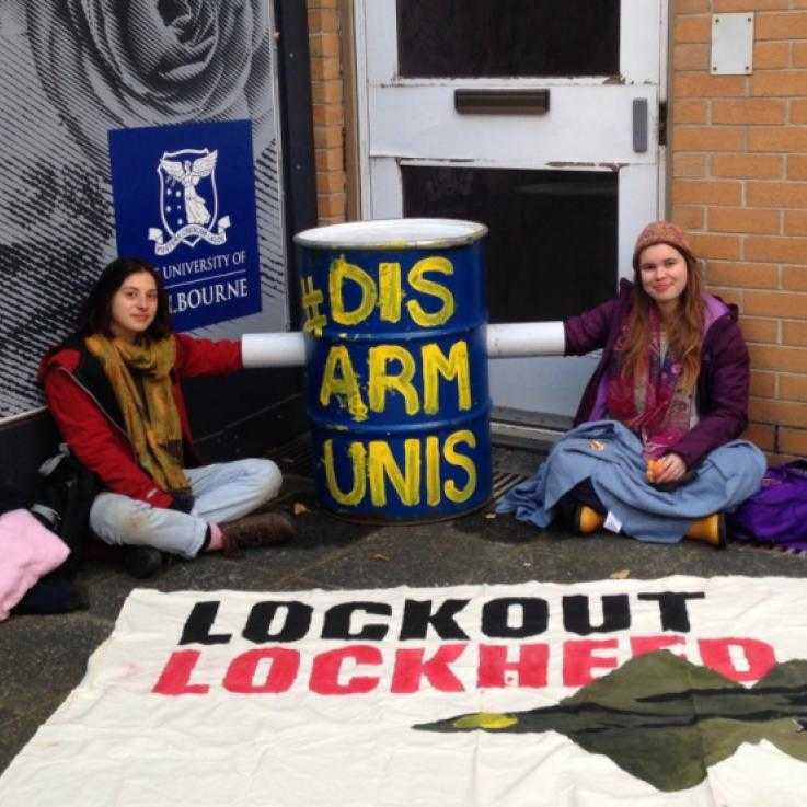 Two student activists lock on to a barrel filled with concrete. The barrel is painted blue and has the words "Disarm Unis" painted on it. In front of the activists there is a banner reading "Lockout Lockheed". Behind them is a sign for the University of Melbourne