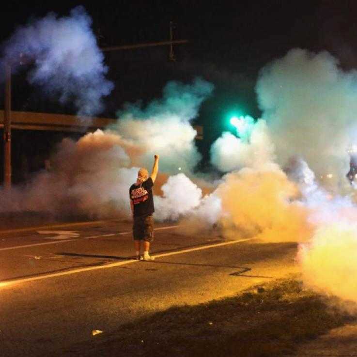 A man stands still with his arm raised amid a cloud of tear gas at night
