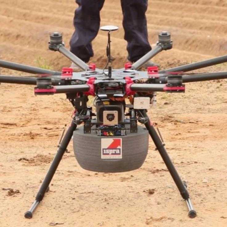 A drone sits on the ground. It has long legs and six arms with rotors. Underneath is a grey box with the word "ISPRA" on the front