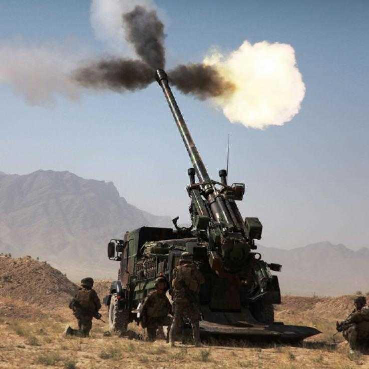 A large cannon is fired in desert terrain. A number of soldiers stand around the side.