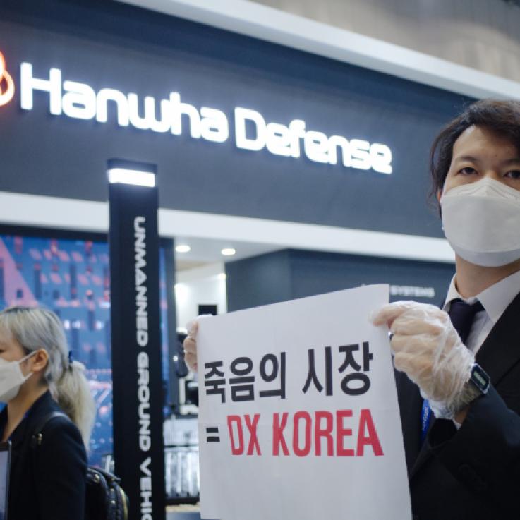 An activist dressed in a suit holds a protest sign in front of an arms exhibitor