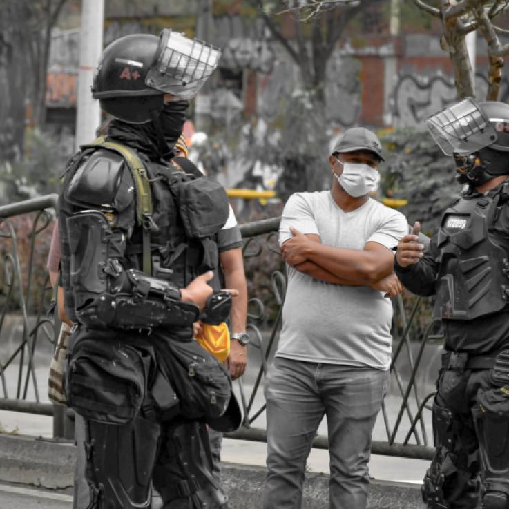 Varios policías fuertemente blindados se sitúan alrededor de un hombre vestido de manera informal. Uno de los policías sostiene una granada